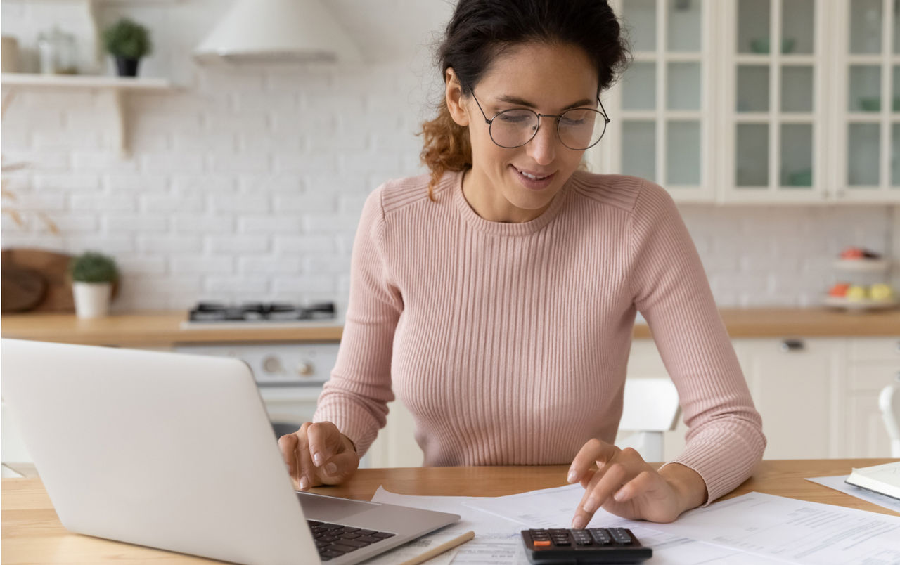 Woman sitting at her kitchen table using her laptop and a calculator